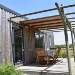 Extérieur d'un lodge premium moderne en bois avec terrasse, pergola et mobilier de jardin face à la nature.