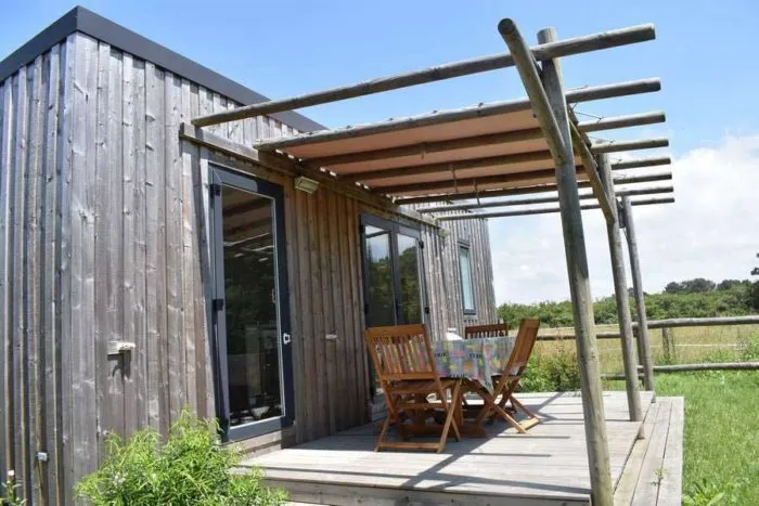 Extérieur d'un lodge premium moderne en bois avec terrasse, pergola et mobilier de jardin face à la nature.